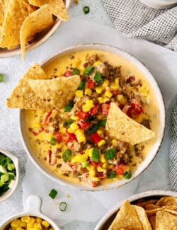 A white bowl of cheesy corn sausage dip is garnished with Rotel and corn chips.