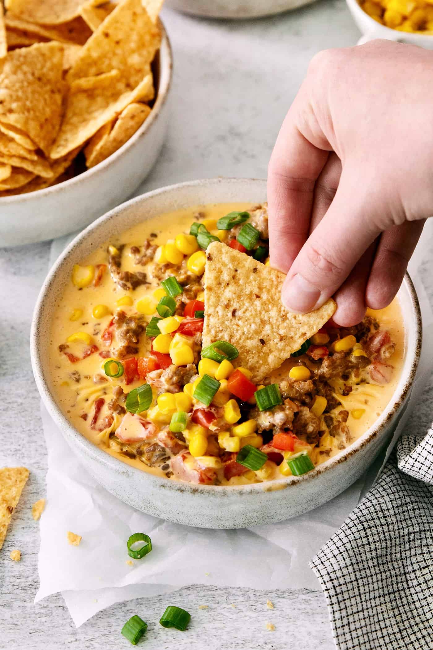 This gorgeously cheesy Sausage Dip takes a creamy base of Velveeta and sour cream and swirls it with spicy sausage and juicy corn kernels. A hand dips a corn chip into a bowl of cheesy corn sausage dip.