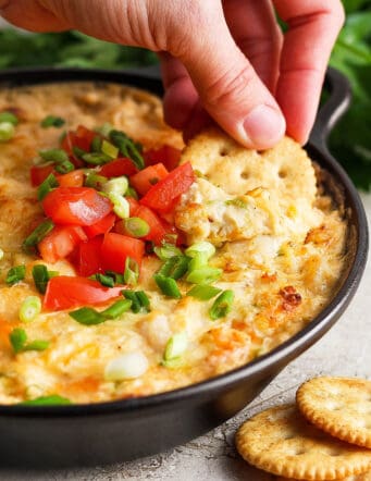 A hand dips a cracker into a tomato-topped bowl of shrimp and crab dip.