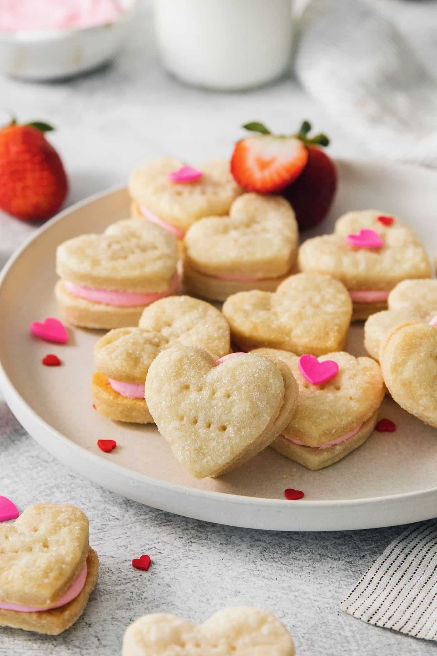 Strawberry Cream Wafer Cookies feature delicate cream and butter cookies that sandwich a pink filling that's fragrant with strawberry extract. Heart-shaped strawberry cream wafer cookies are displayed on a white plate.