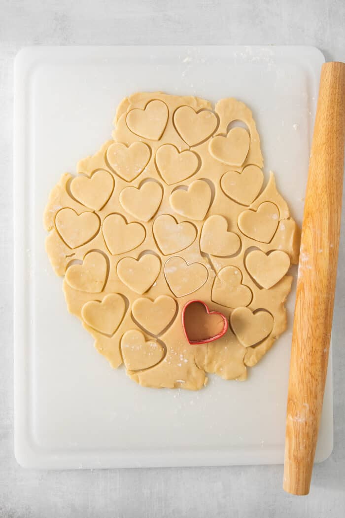 Strawberry Cream Wafer Cookies feature delicate cream and butter cookies that sandwich a pink filling that's fragrant with strawberry extract. Heart shapes are cut out of a piece of cream wafer cookie dough on a white cutting board, with a rolling pin to the right.