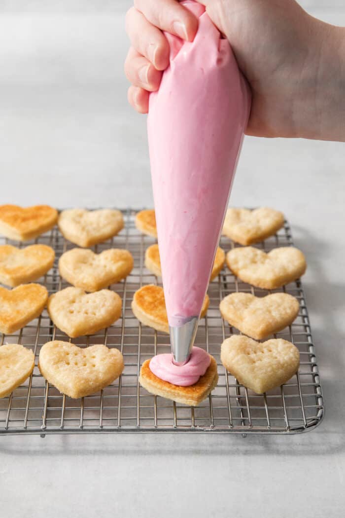 Strawberry Cream Wafer Cookies feature delicate cream and butter cookies that sandwich a pink filling that's fragrant with strawberry extract. Strawberry buttercream is piped onto heart shaped cream wafer cookies on a wire rack.