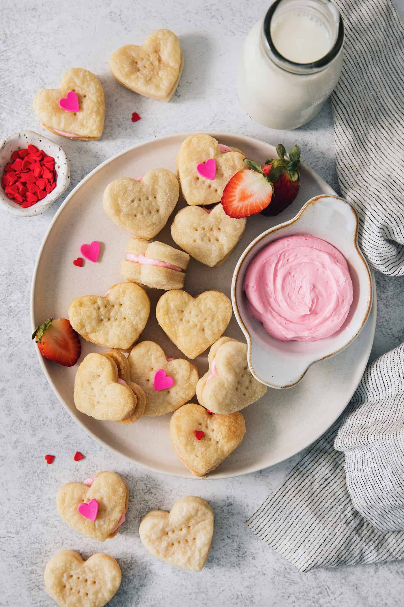 Strawberry Cream Wafer Cookies feature delicate cream and butter cookies that sandwich a pink filling that's fragrant with strawberry extract. A serving dish holds strawberry cream wafer cookies decorated with heart sprinkles, with a bowl of strawberry buttercream next o them.