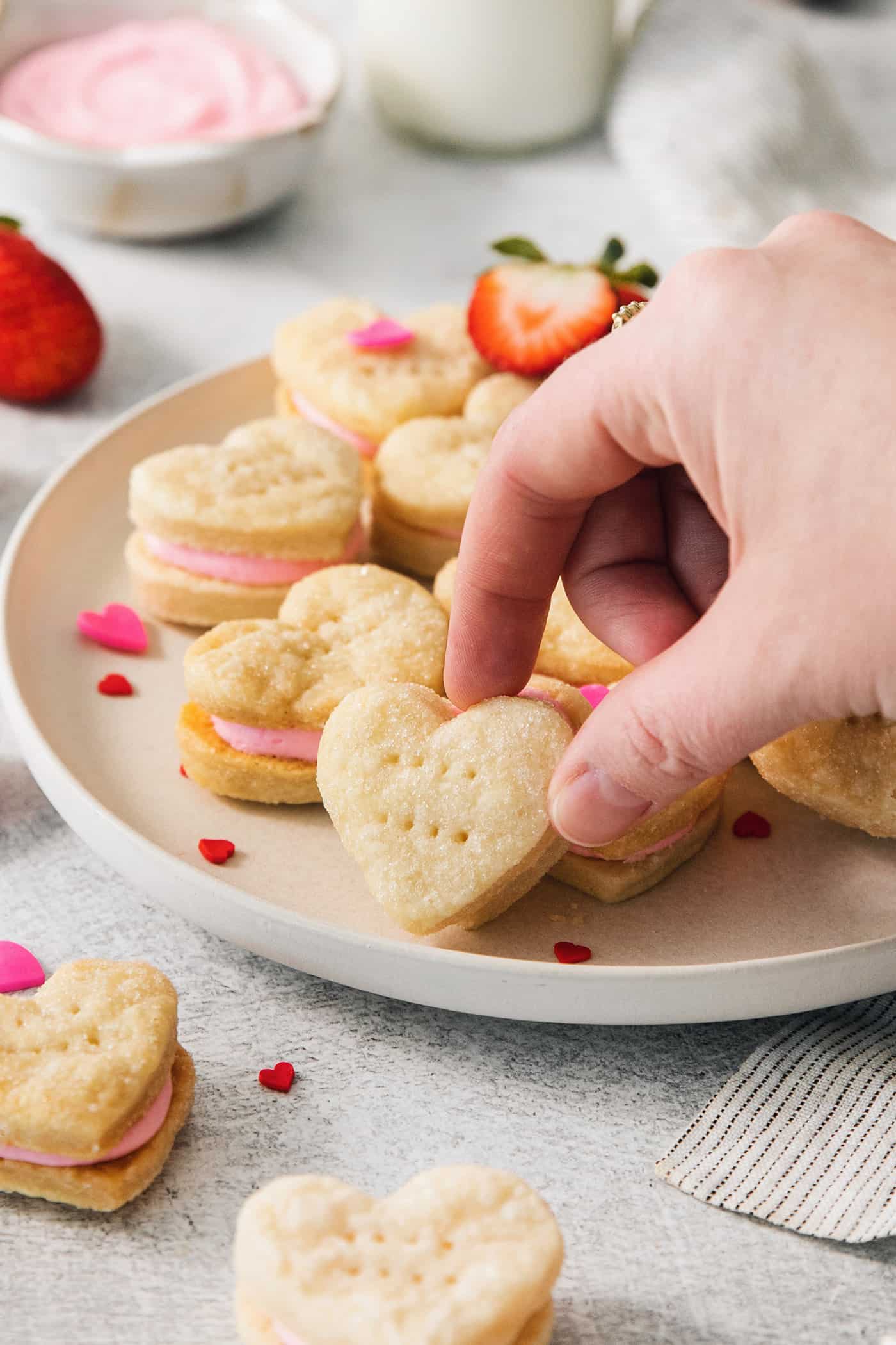 Strawberry Cream Wafer Cookies feature delicate cream and butter cookies that sandwich a pink filling that's fragrant with strawberry extract. A hand reaches to pick up a heart shaped strawberry cream wafter cookie from a plate of cookies.