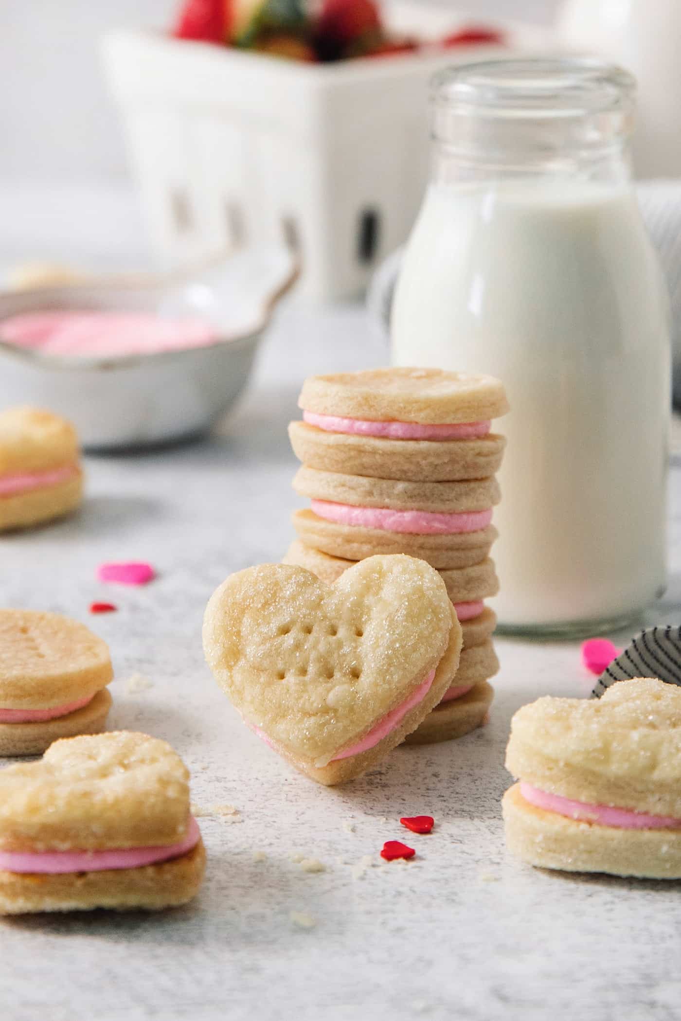 Strawberry Cream Wafer Cookies feature delicate cream and butter cookies that sandwich a pink filling that's fragrant with strawberry extract. A stack of strawberry cream wafter cookies is surrounded by more cookies, with a jug of milk and a bowl of pink buttercream in the background.