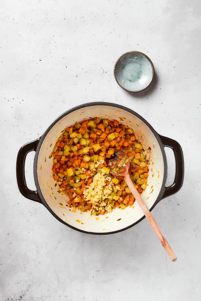 Greek-inspired Lemon Chicken Orzo Soup is fragrant with garlic and packed with tender chicken, vegetables, and delicate orzo pasta. A soup base including onions, carrots, and celery cooks in a pot with a wooden spoon to stir.