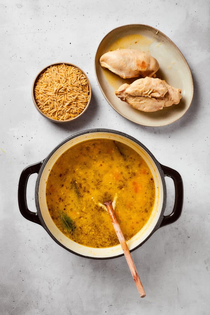 Greek-inspired Lemon Chicken Orzo Soup is fragrant with garlic and packed with tender chicken, vegetables, and delicate orzo pasta. The cooked chicken is removed from the pot and placed alongside it, with a bowl of orzo waiting to be added.