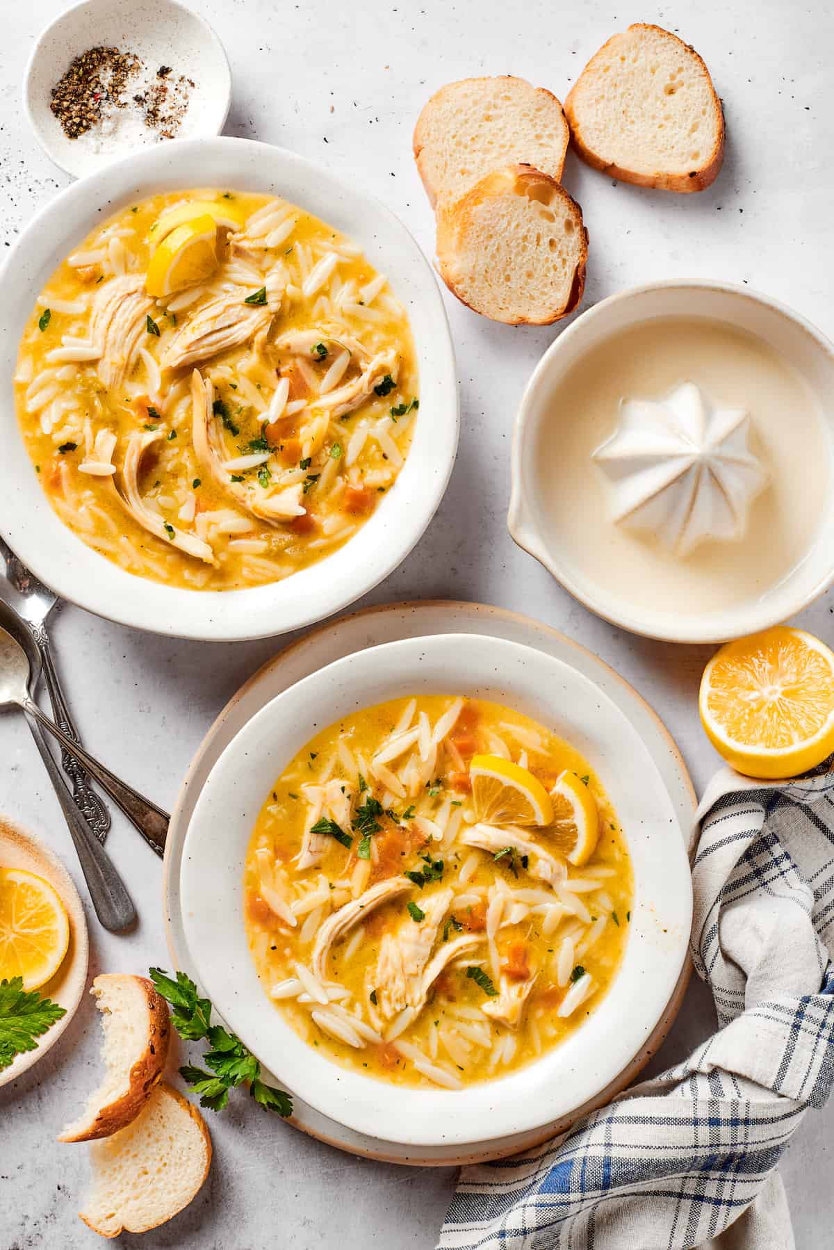 Greek-inspired Lemon Chicken Orzo Soup is fragrant with garlic and packed with tender chicken, vegetables, and delicate orzo pasta. Bowls of lemon chicken orzo soup on a table with a juicer and lemon halves nearby.