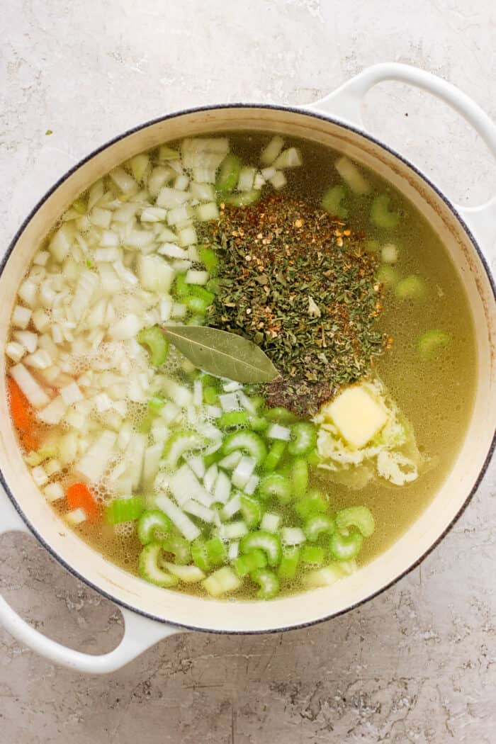 This homemade Chicken Noodle Soup is an easy classic with from-scratch chicken broth loaded with tender chicken, vegetables, and egg noodles. Chopped carrots, onion, celery, garlic, and herbs are shown on top of a pot of chicken broth.
