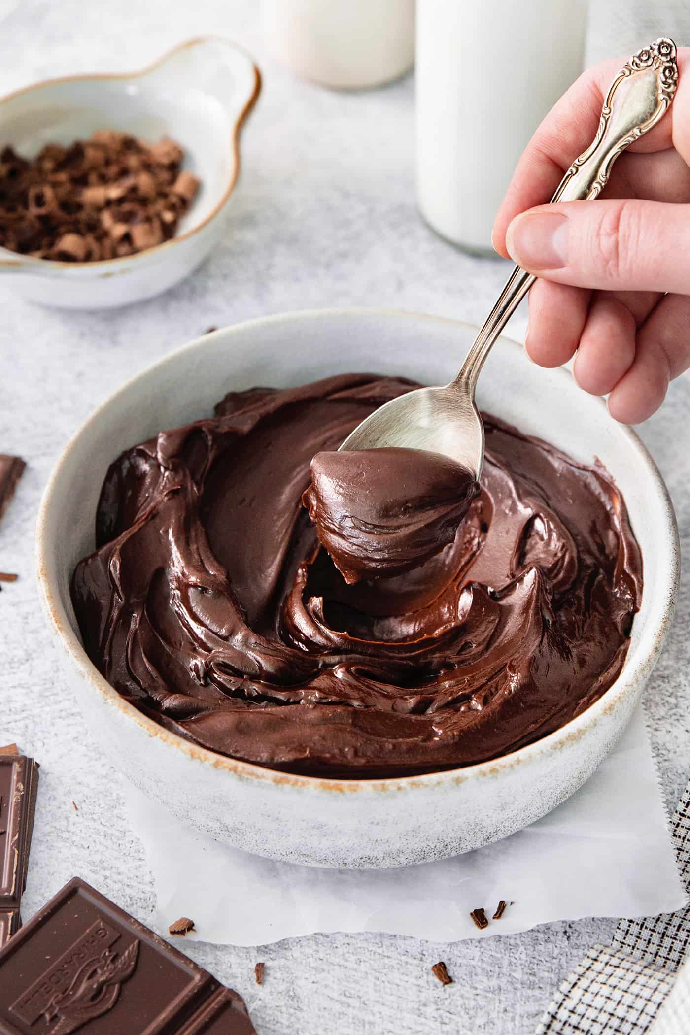 This easy Chocolate Ganache recipe is a versatile staple, perfect for so many things – from cakes to tart fillings to drizzling over coffee! A hand holding a silver spoon stirs a white bowl of chocolate ganache.