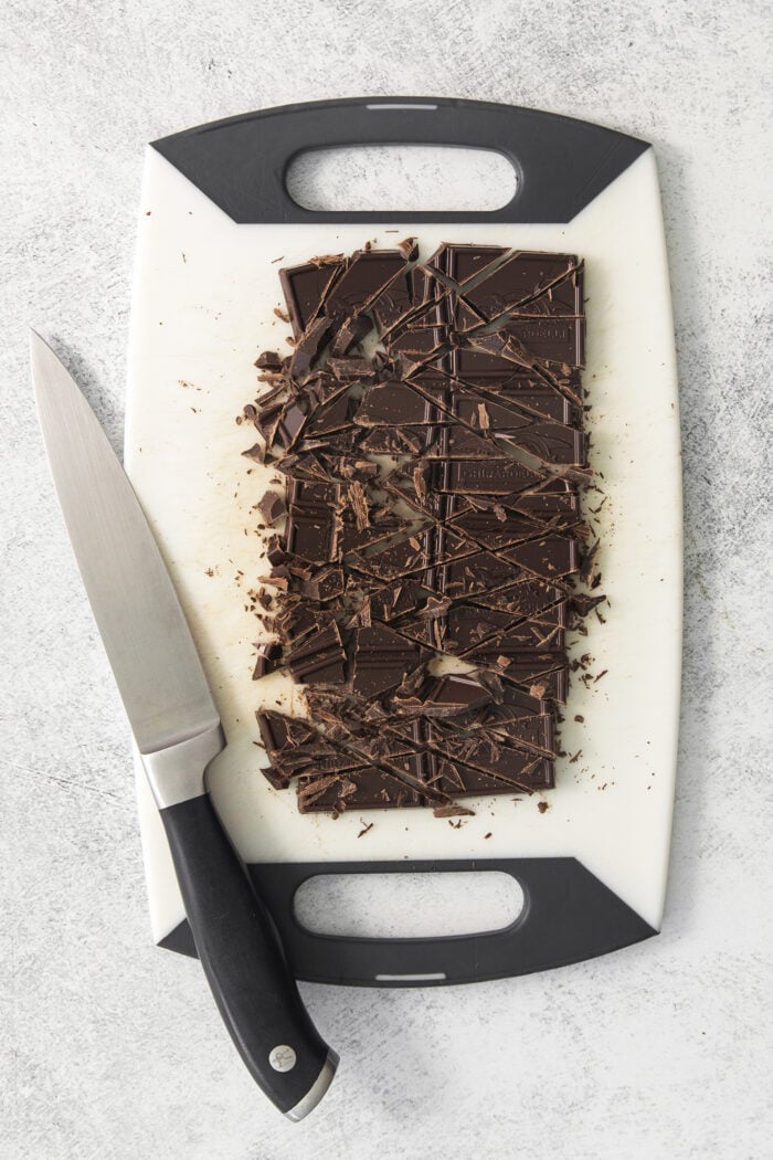 This easy Chocolate Ganache recipe is a versatile staple, perfect for so many things – from cakes to tart fillings to drizzling over coffee! A white cutting board holds chopped chocolate with a knif resting on its left edge.