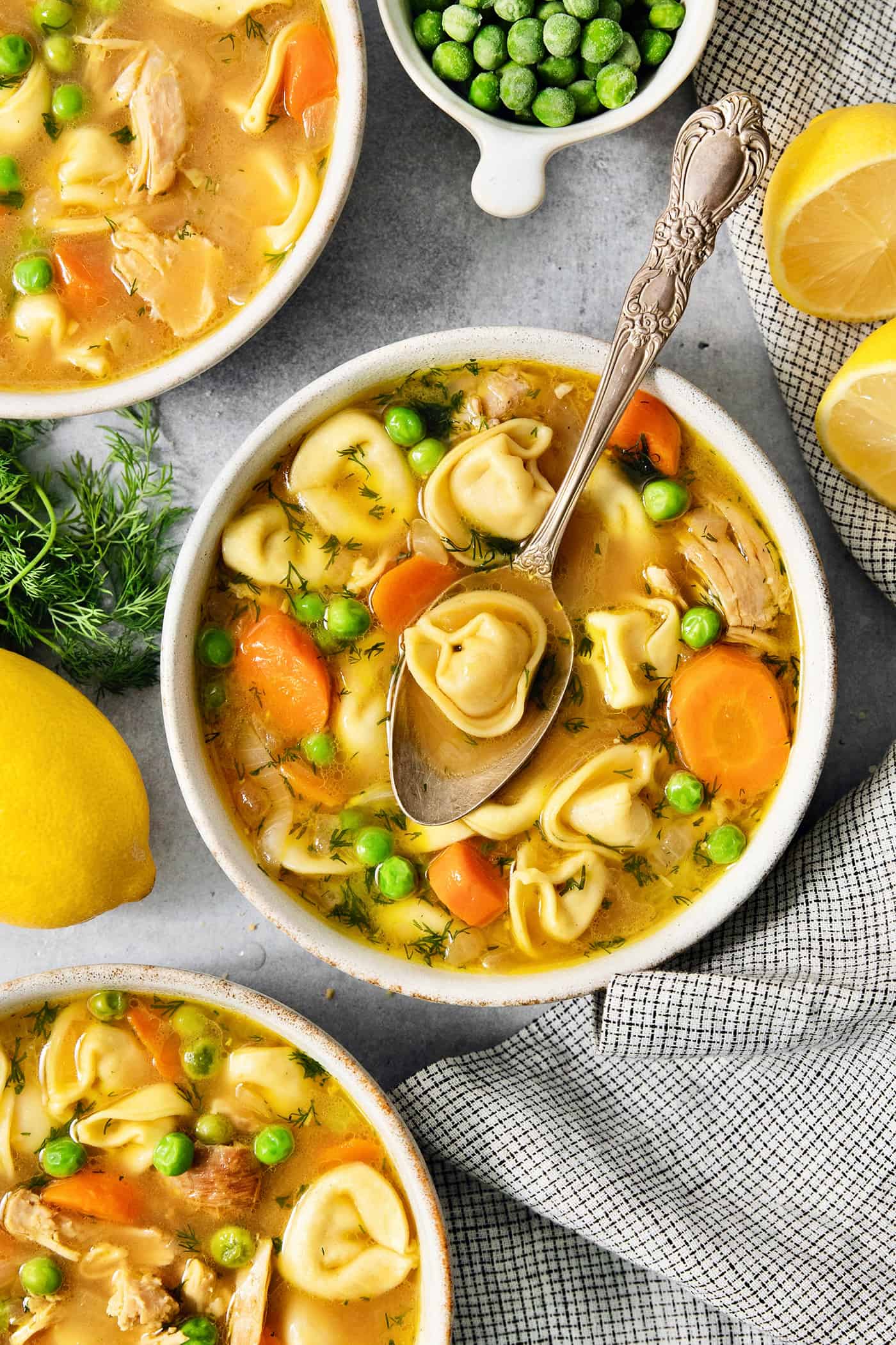 Cheesy tortellini pockets are the stars of this Lemon Chicken Tortellini Soup that also features a lemony broth packed with shredded chicken. A bowl of lemon chicken tortellini soup on a table surrounded by more bowls of soup, peas, and lemons.