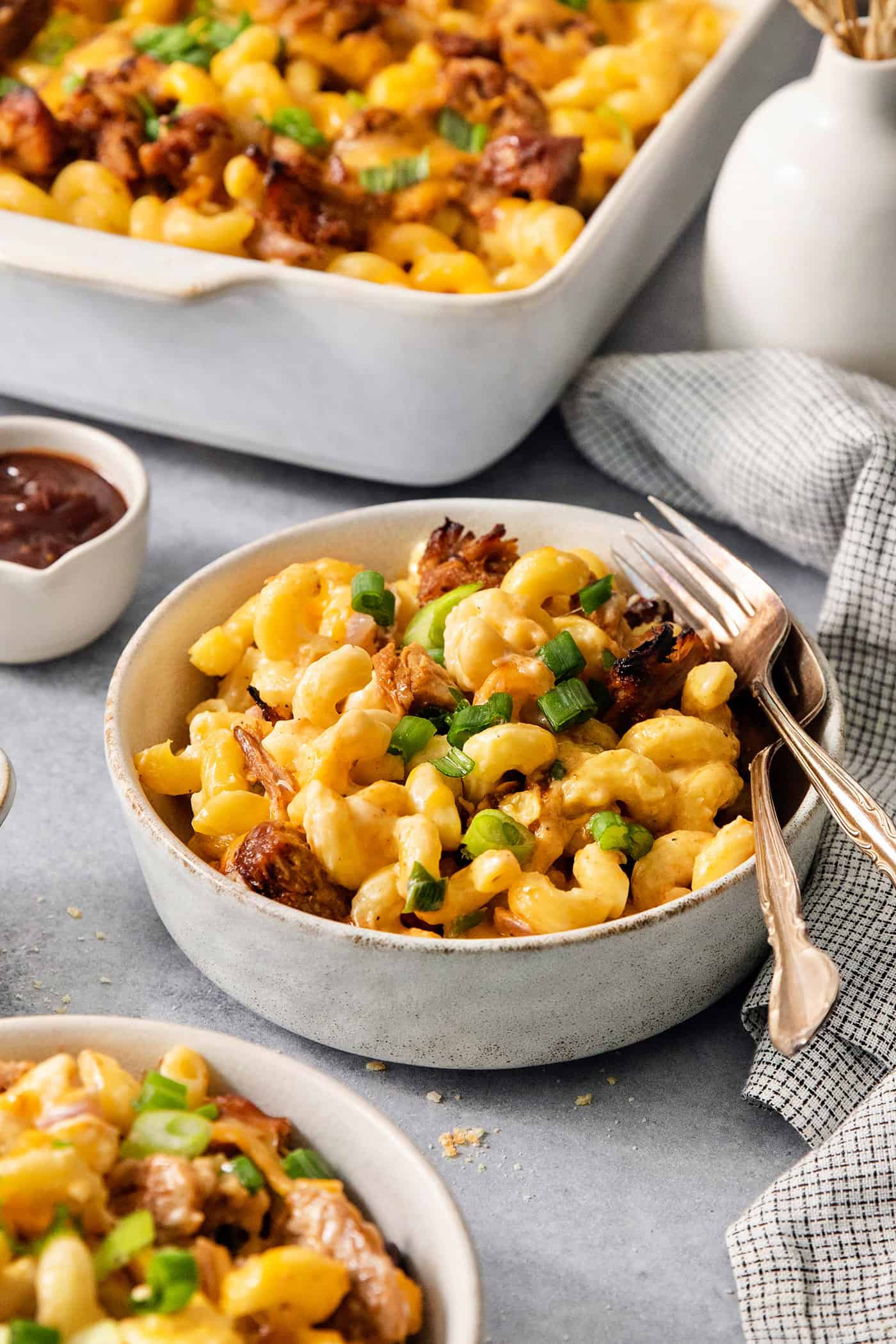 This easy Pulled Pork Mac and Cheese is super decadent with rich and creamy 3-cheese macaroni and cheese plus tender shredded pork. Bowls of pulled pork and cheese on a table with a pan of mac and cheese in the background.