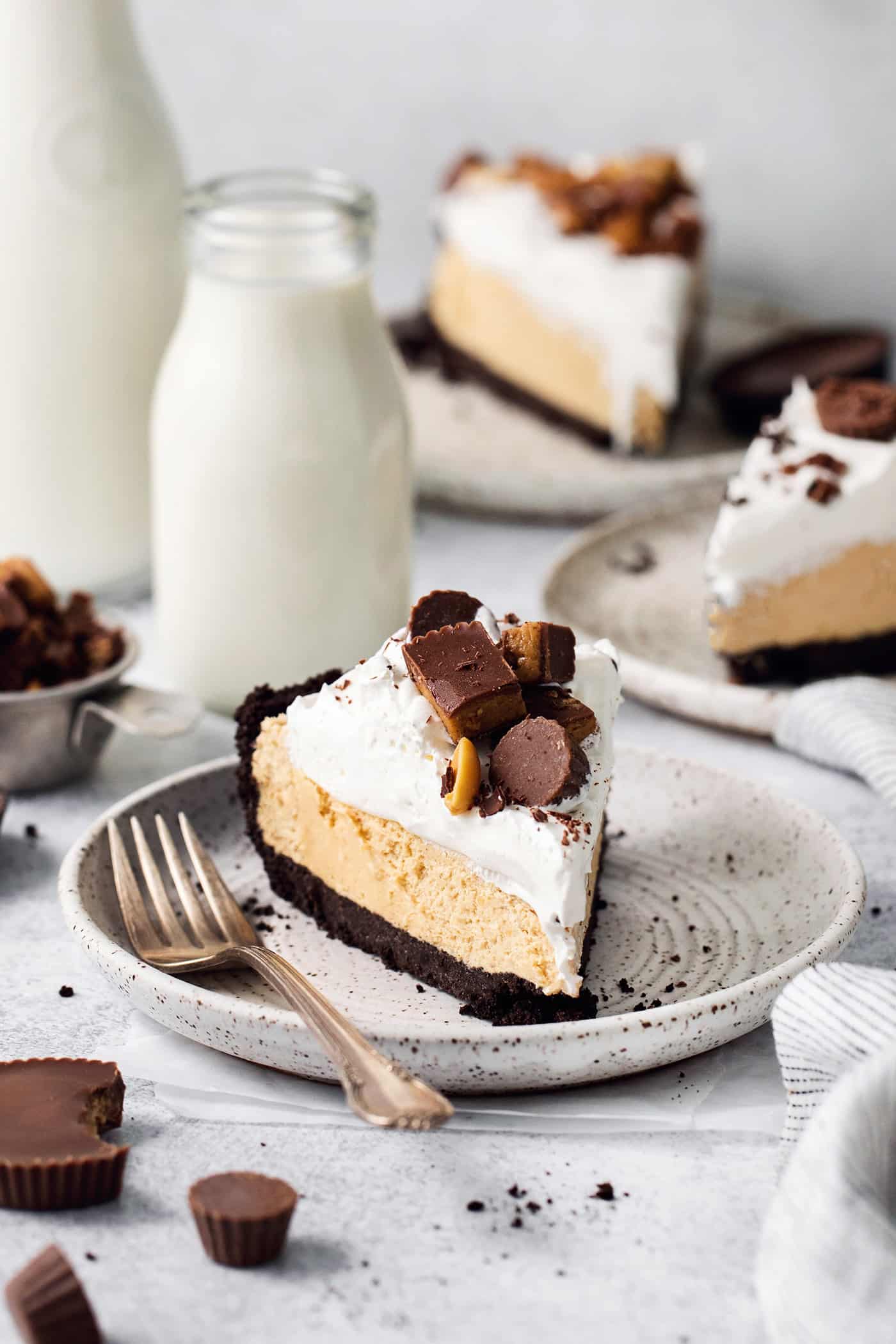 This no-bake Reese’s Pie features a crisp Oreo crust and a creamy peanut butter filling finished with whipped topping and Reese’s cups. A candy-topped piece of Reeses pie is shown on a plate with more slices of pie around it.