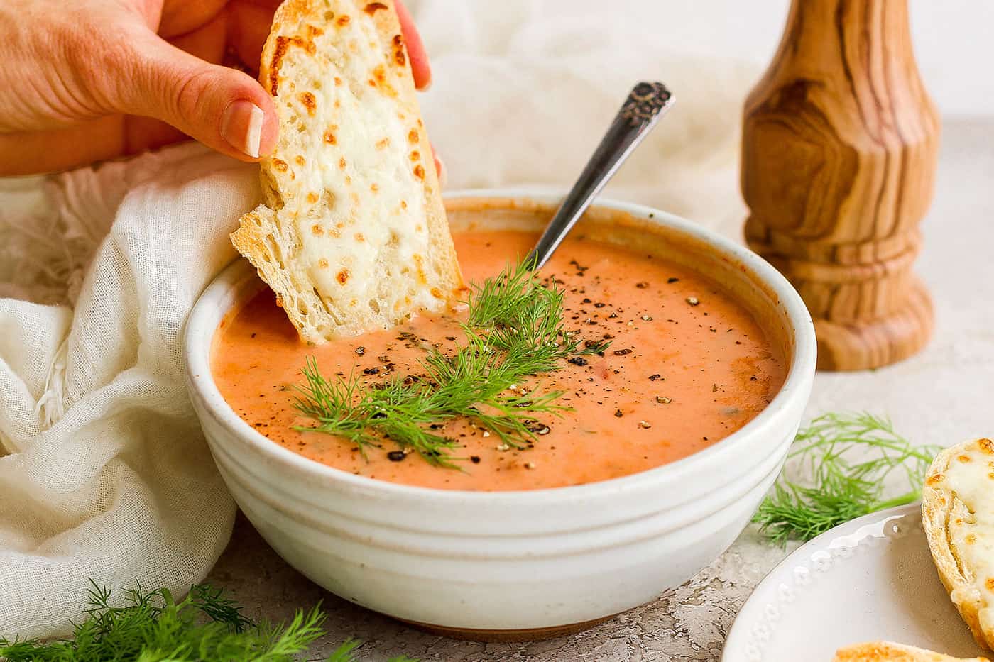 a hand dipping a piece of cheesy bread into a bowl of creamy tomato soup