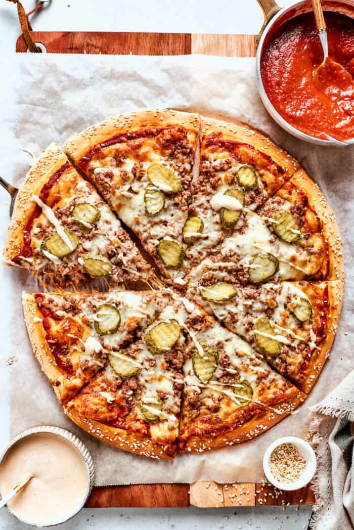 This Cheeseburger Pizza recipe turns your favorite burger into pizza! Top homemade dough with pizza sauce, ground beef, cheese, and pickles. Bowls of sauce are arranged around a cheeseburger pizza.