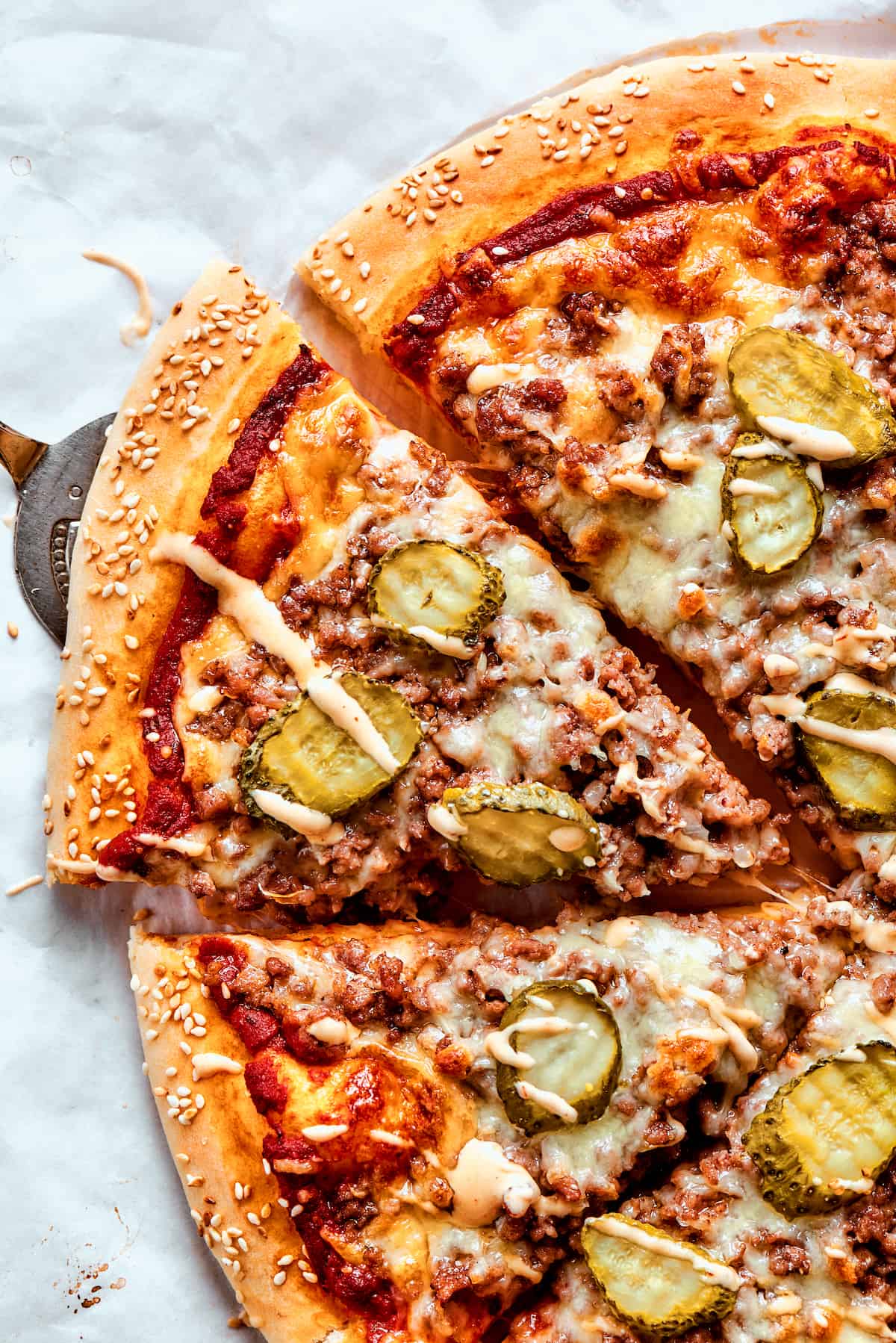 This Cheeseburger Pizza recipe turns your favorite burger into pizza! Top homemade dough with pizza sauce, ground beef, cheese, and pickles. Half of a sliced cheeseburger pizza on a white background.