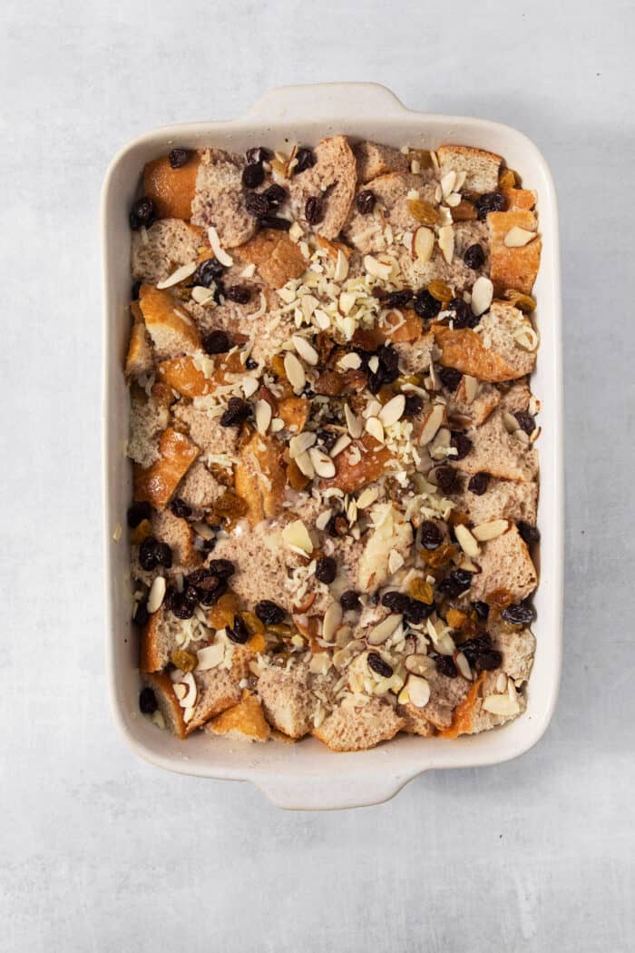 Capirotada, or Mexican Bread Pudding, features layers of crusty bread, raisins, sliced almonds, and cheese soaked in spiced sweet cream. A baking dish of capirotada is shown before being placed in the oven.