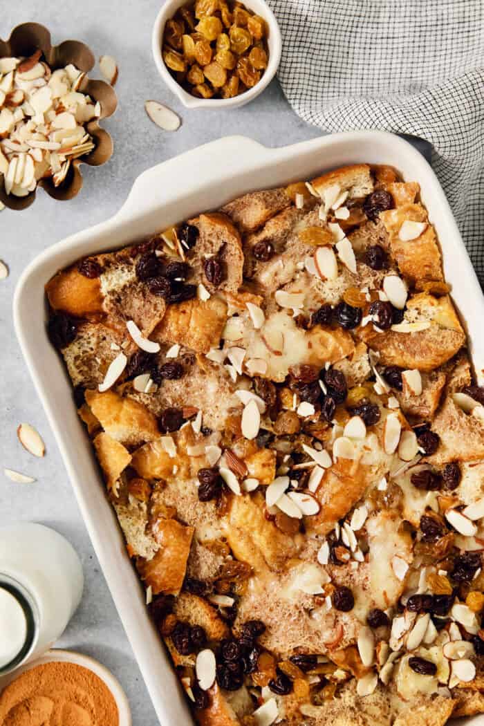 Capirotada, or Mexican Bread Pudding, features layers of crusty bread, raisins, sliced almonds, and cheese soaked in spiced sweet cream. A baking dish of capirotada is surrounded by bowls of almonds and raisins.