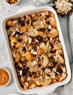 Bowls of golden raisins and almonds surround a white baking dish full of capirotada.