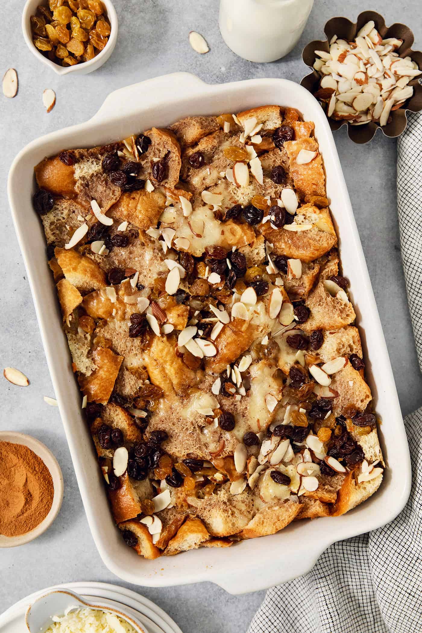 Capirotada, or Mexican Bread Pudding, features layers of crusty bread, raisins, sliced almonds, and cheese soaked in spiced sweet cream. Bowls of golden raisins and almonds surround a white baking dish full of capirotada.