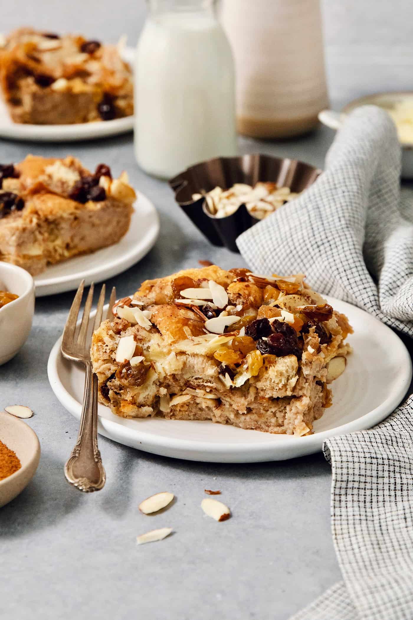 Capirotada, or Mexican Bread Pudding, features layers of crusty bread, raisins, sliced almonds, and cheese soaked in spiced sweet cream. A portion of capirotada is served on a white plate with a fork.