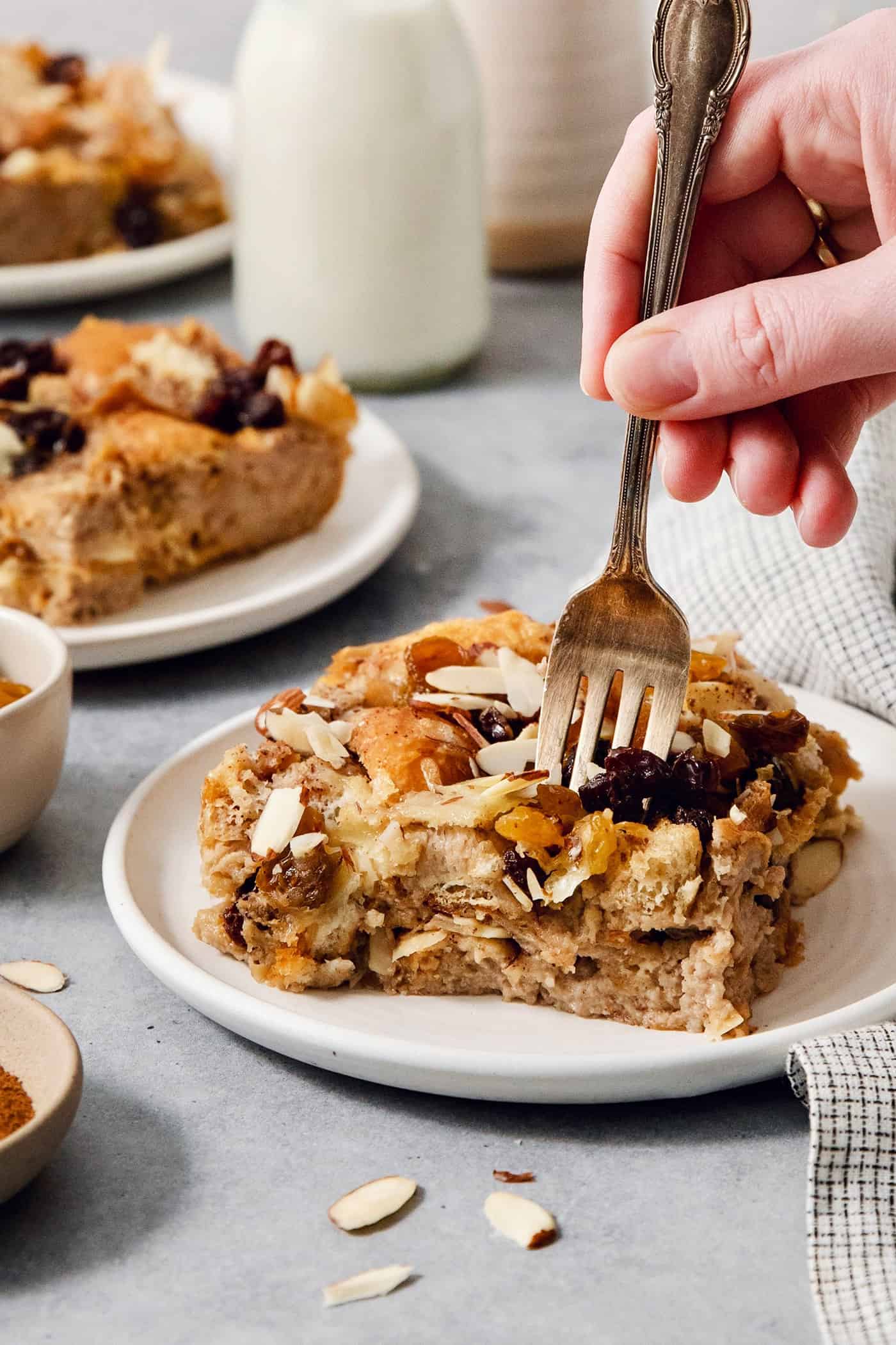 Capirotada, or Mexican Bread Pudding, features layers of crusty bread, raisins, sliced almonds, and cheese soaked in spiced sweet cream. A hand holding a fork cuts into a portion of capirotada on a white plate.