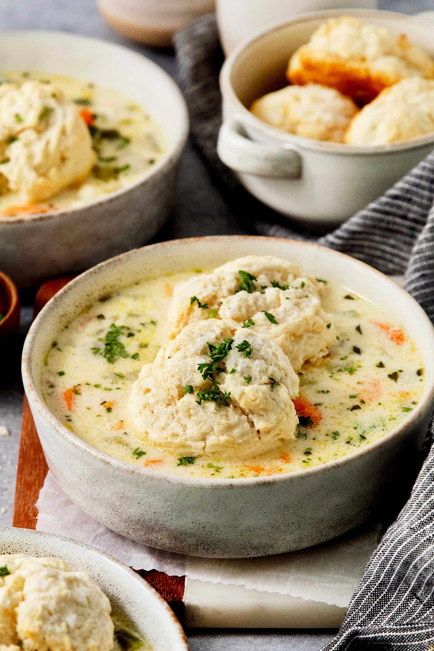 This homemade Chicken Pot Pie Soup recipe features a creamy broth that’s loaded with chicken and vegetables and topped with biscuits. Bowls of biscuit-topped chicken pot pie soup with a basket of biscuits behind them.