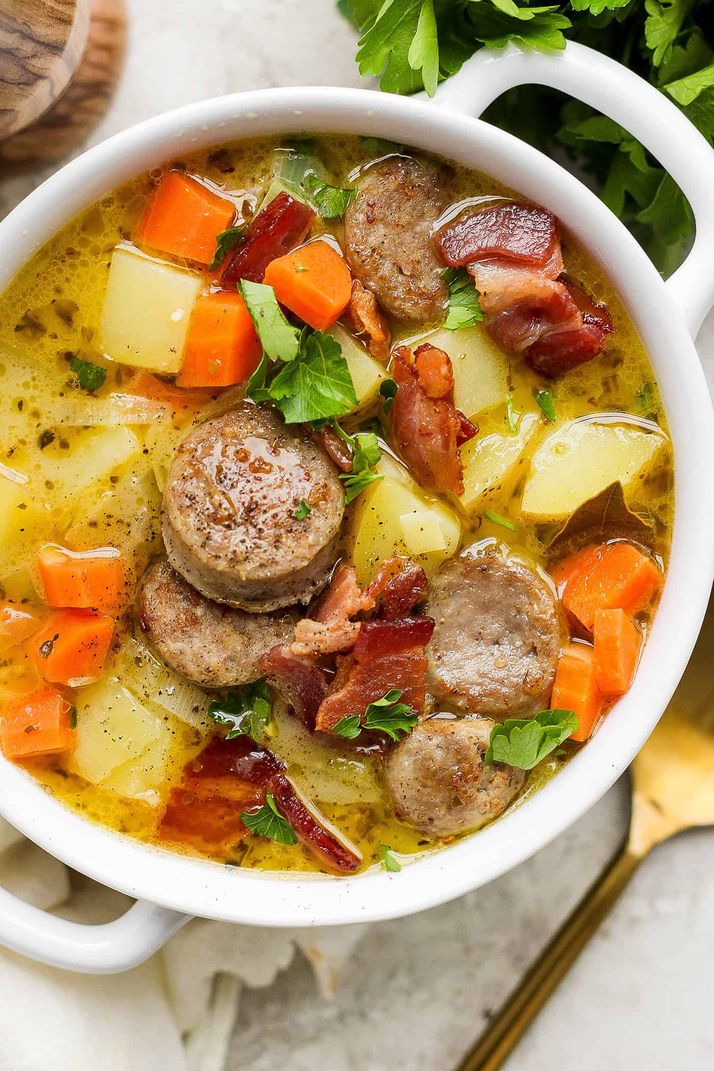 This German Potato Soup recipe is packed with savory sausage, salty bacon, and plenty of vegetables plus a spoonful of half-and-half. A white handled bowl with a serving of German potato soup.