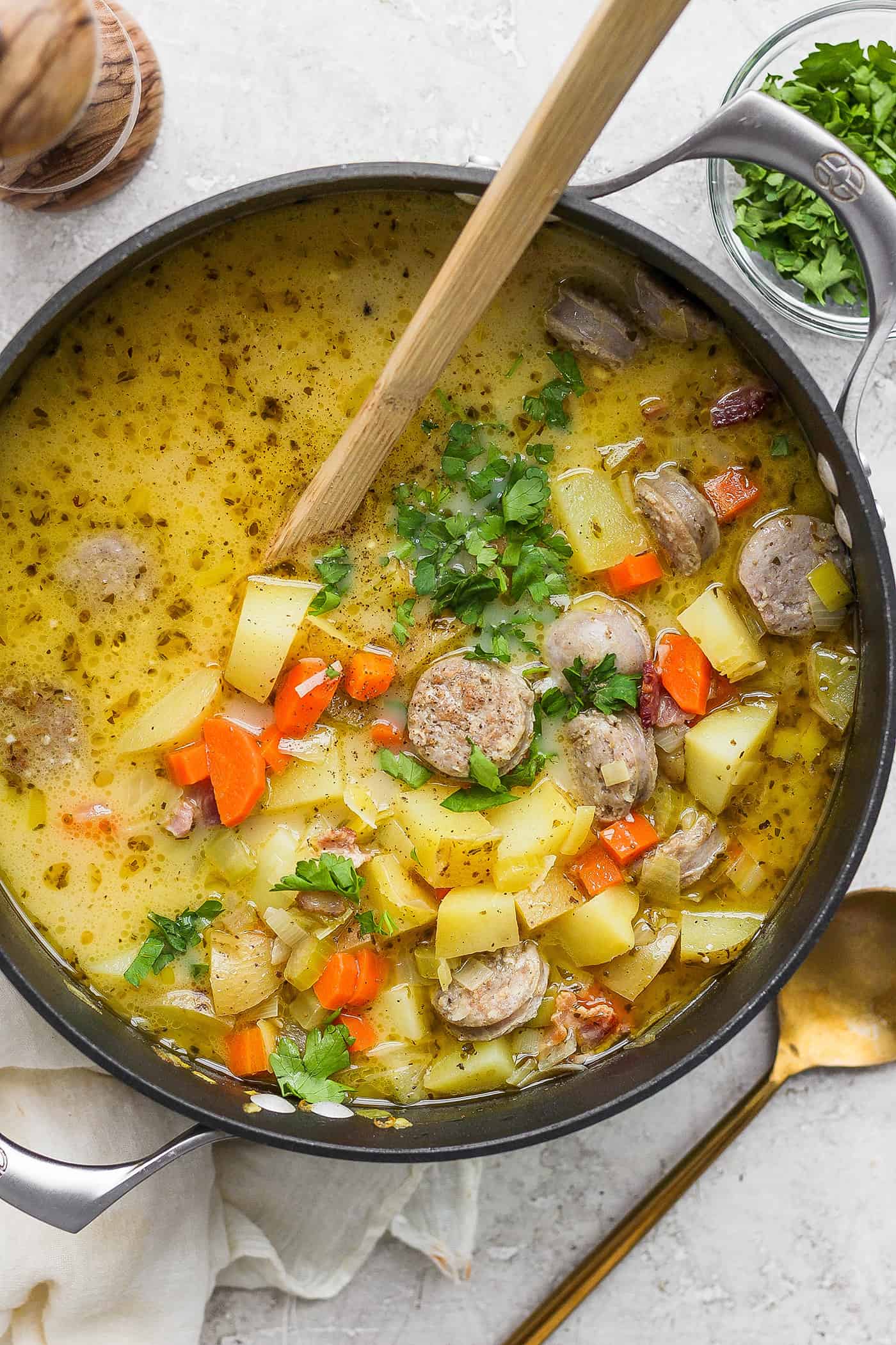 This German Potato Soup recipe is packed with savory sausage, salty bacon, and plenty of vegetables plus a spoonful of half-and-half. A wooden spoon stirs a pot of German potato soup.