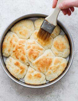 A hand uses a pastry brush to brush the baked biscuits with melted butter.