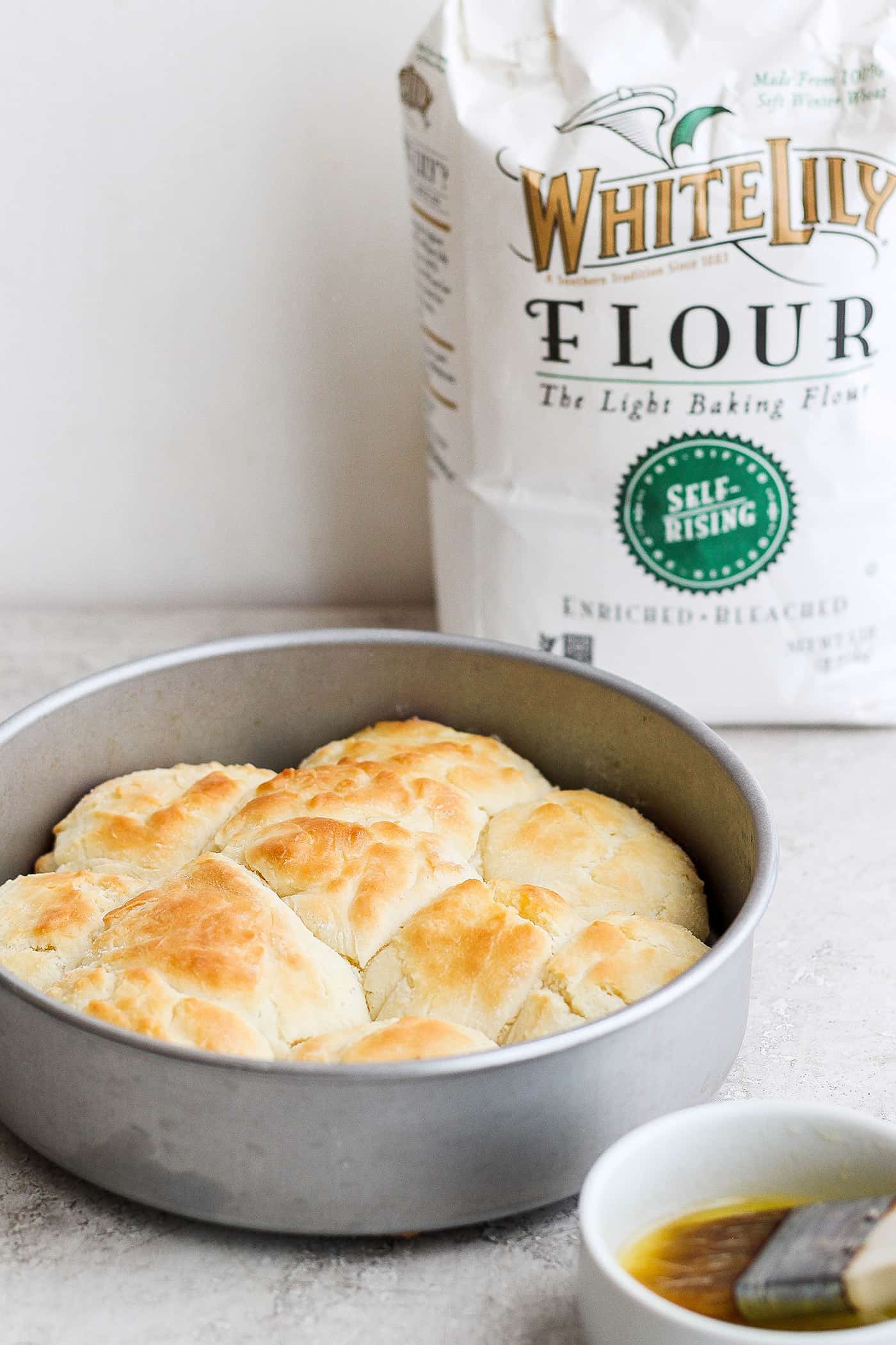Flaky and light Touch of Grace Biscuits are made with White Lily flour to make unbelievably tender pull-apart biscuits. A bag of White Lily flour is seen in the background with baked touch of grace biscuits in a baking tin in the foreground.