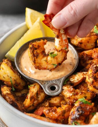 A hand dips a piece of blackened shrimp into a bowl of sauce.