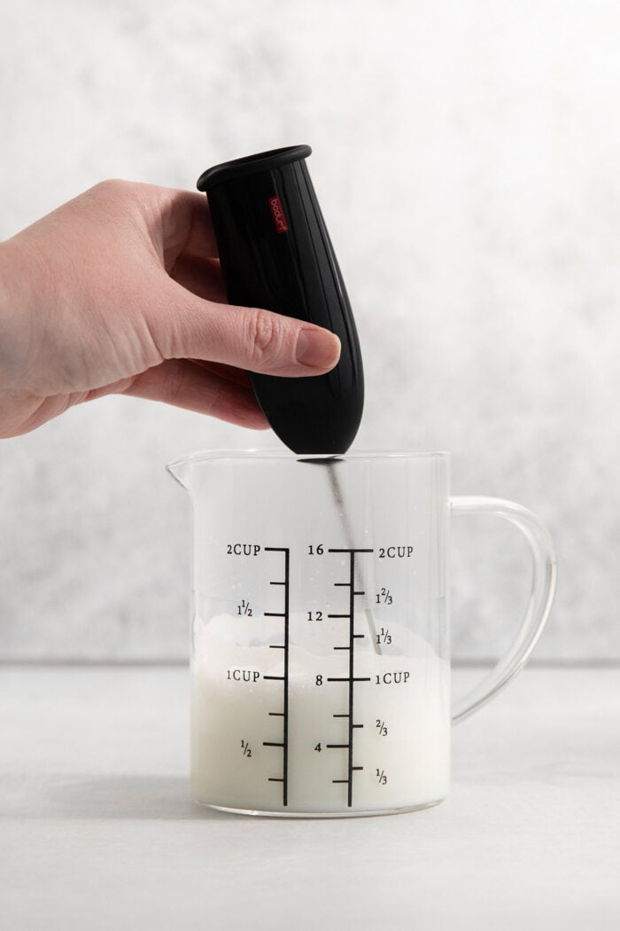 This Lavender Latte mixes freshly brewed espresso, oat milk, and a sweet lavender simple syrup to create a copycat Starbucks lavender latte. A hand holds a milk frother to froth oat milk in a glass pitcher.