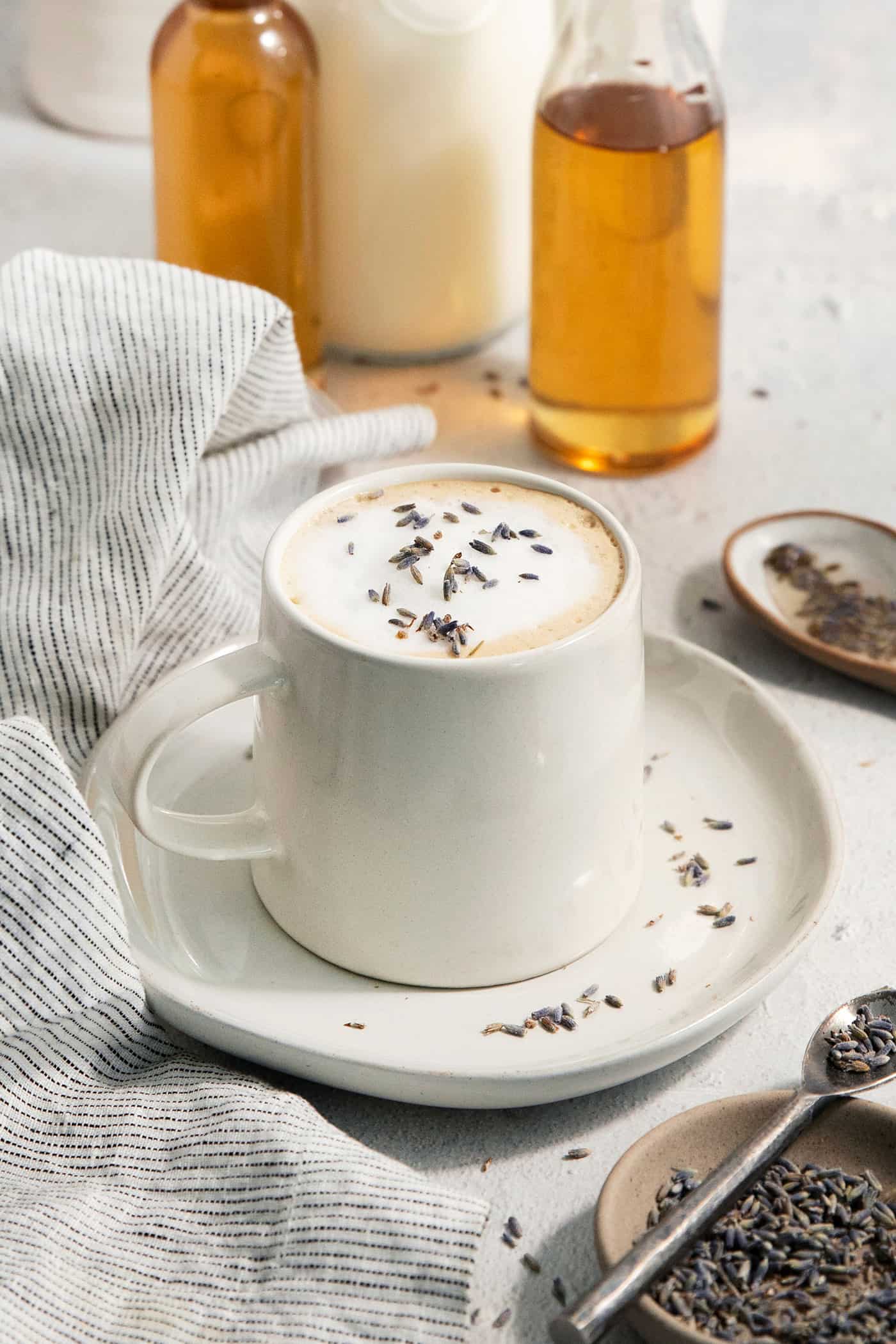 This Lavender Latte mixes freshly brewed espresso, oat milk, and a sweet lavender simple syrup to create a copycat Starbucks lavender latte. A mug of lavender latte on a plate with lavender simple syrup in a jar in the background.