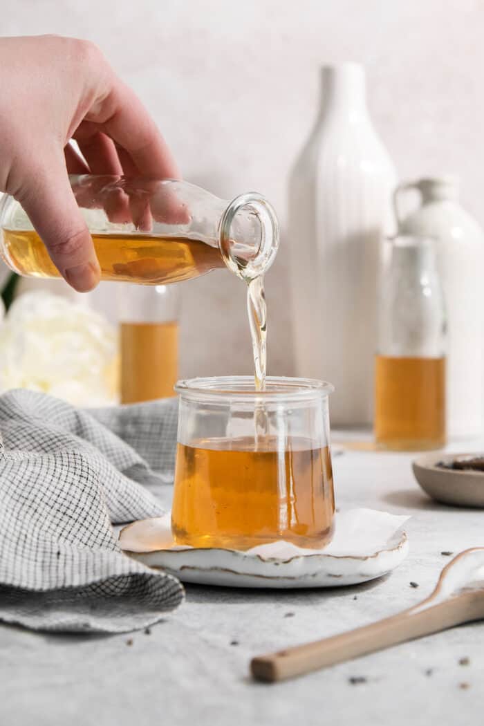 Floral lavender infuses this Lavender Simple Syrup with a hint of sweetness and fresh flavor. Swirl it into coffee and add it to cocktails. A hand pours lavender simple syrup from a bottle into a jar.
