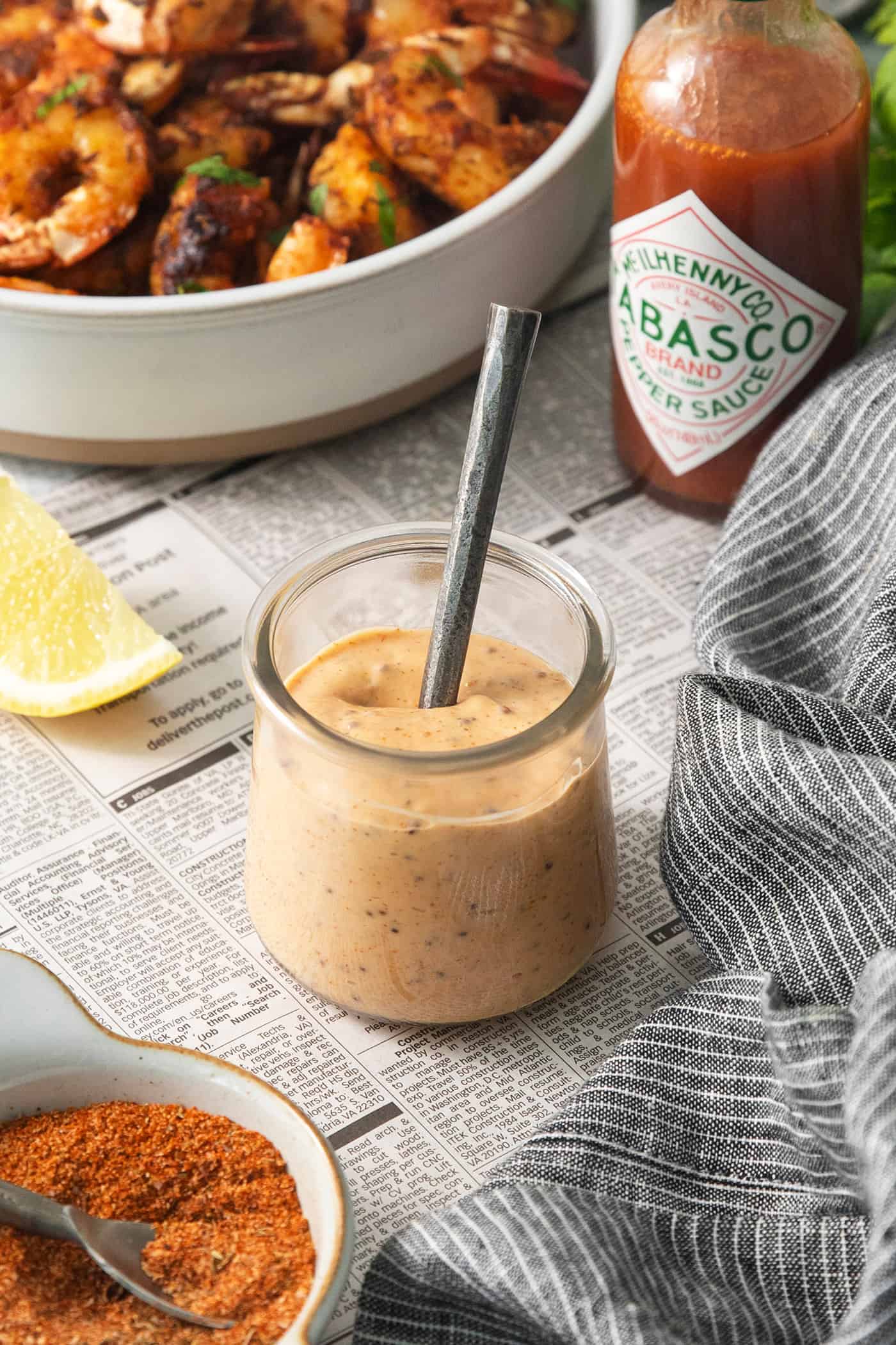 A jar of shrimp dipping sauce with a spoon in it shown in front of a bowl of blackened shrimp.
