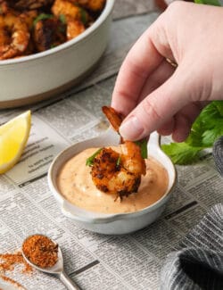 A hand dips a piece of blackened shrimp into a bowl of shrimp dipping sauce.