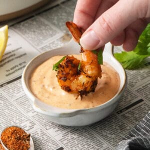 A hand dips a piece of blackened shrimp into a bowl of shrimp dipping sauce.