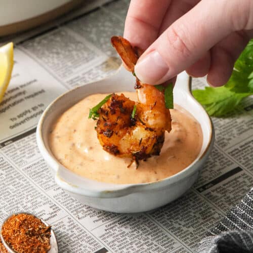 A hand dips a piece of blackened shrimp into a bowl of shrimp dipping sauce.