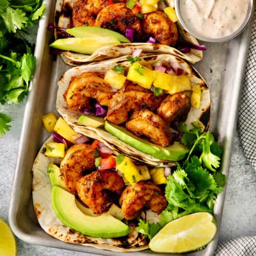 A trays holds blackened shrimp tacos surrounded by lime wedges and a bowls of avocado crema.