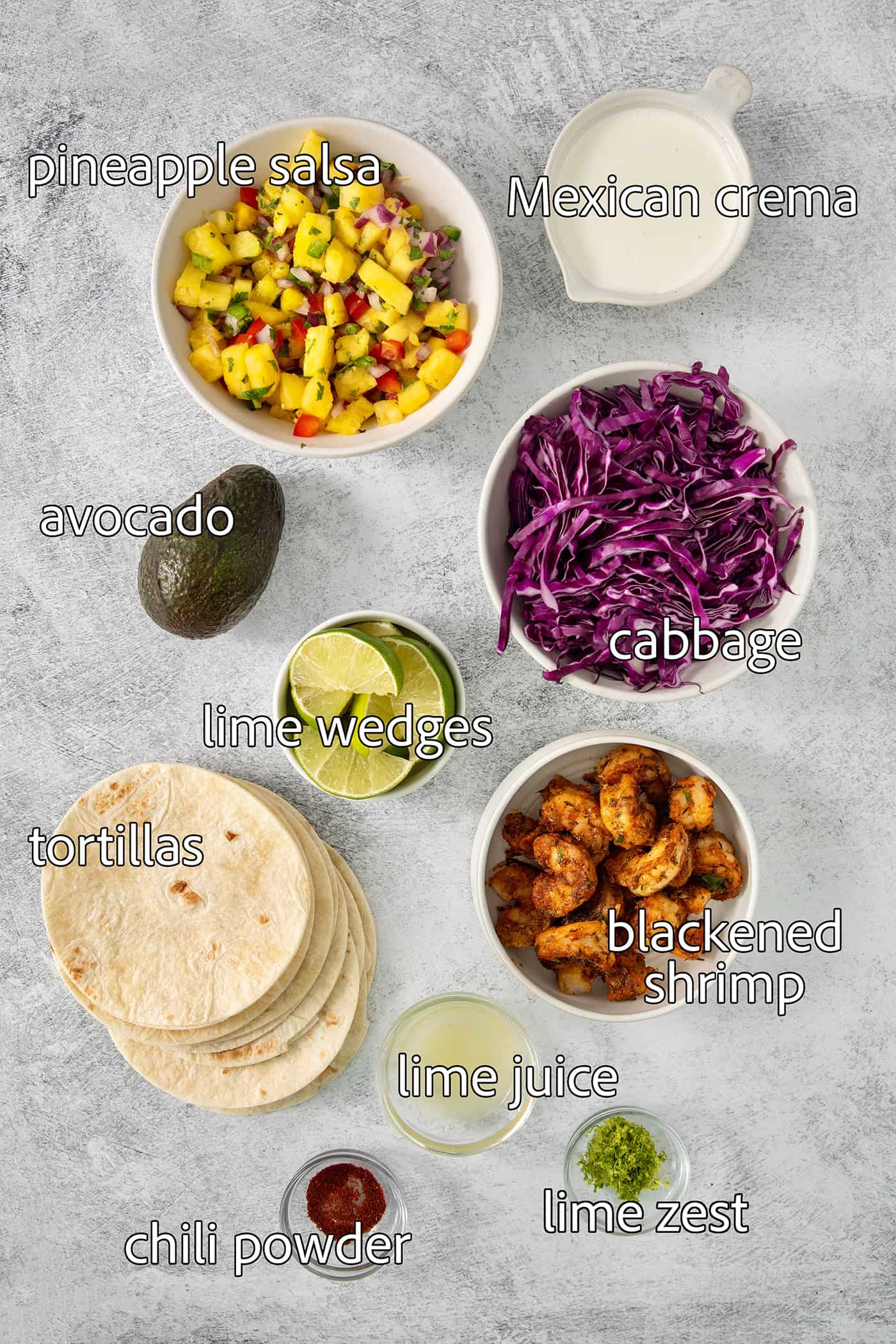These blackened shrimp tacos feature charred flour tortillas stuffed with spicy blackened shrimp, pineapple salsa, and refreshing lime crema. The ingredients for blackened shrimp tacos are portioned out and labeled: blackened shrimp, pineapple salsa, tortillas, cabbage, avocado, crema, lime.