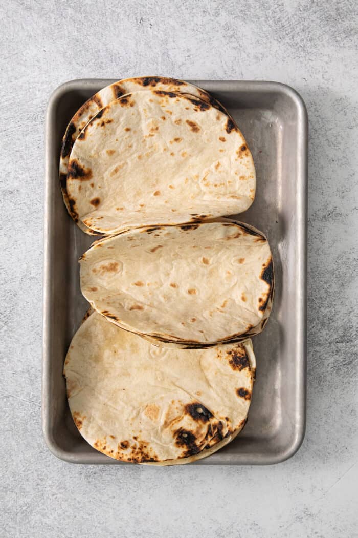 These blackened shrimp tacos feature charred flour tortillas stuffed with spicy blackened shrimp, pineapple salsa, and refreshing lime crema. Charred tortillas are placed on a metal baking pan.