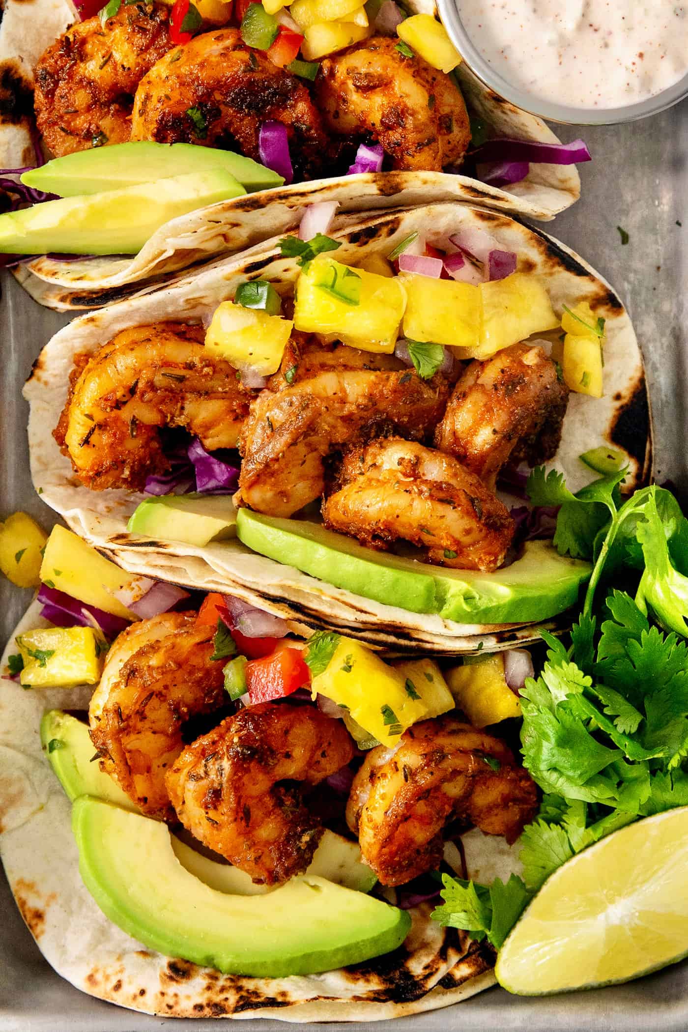 These blackened shrimp tacos feature charred flour tortillas stuffed with spicy blackened shrimp, pineapple salsa, and refreshing lime crema. A close up shows pieces of blackened shrimp in tortillas with avocado slices and pineapple chunks.