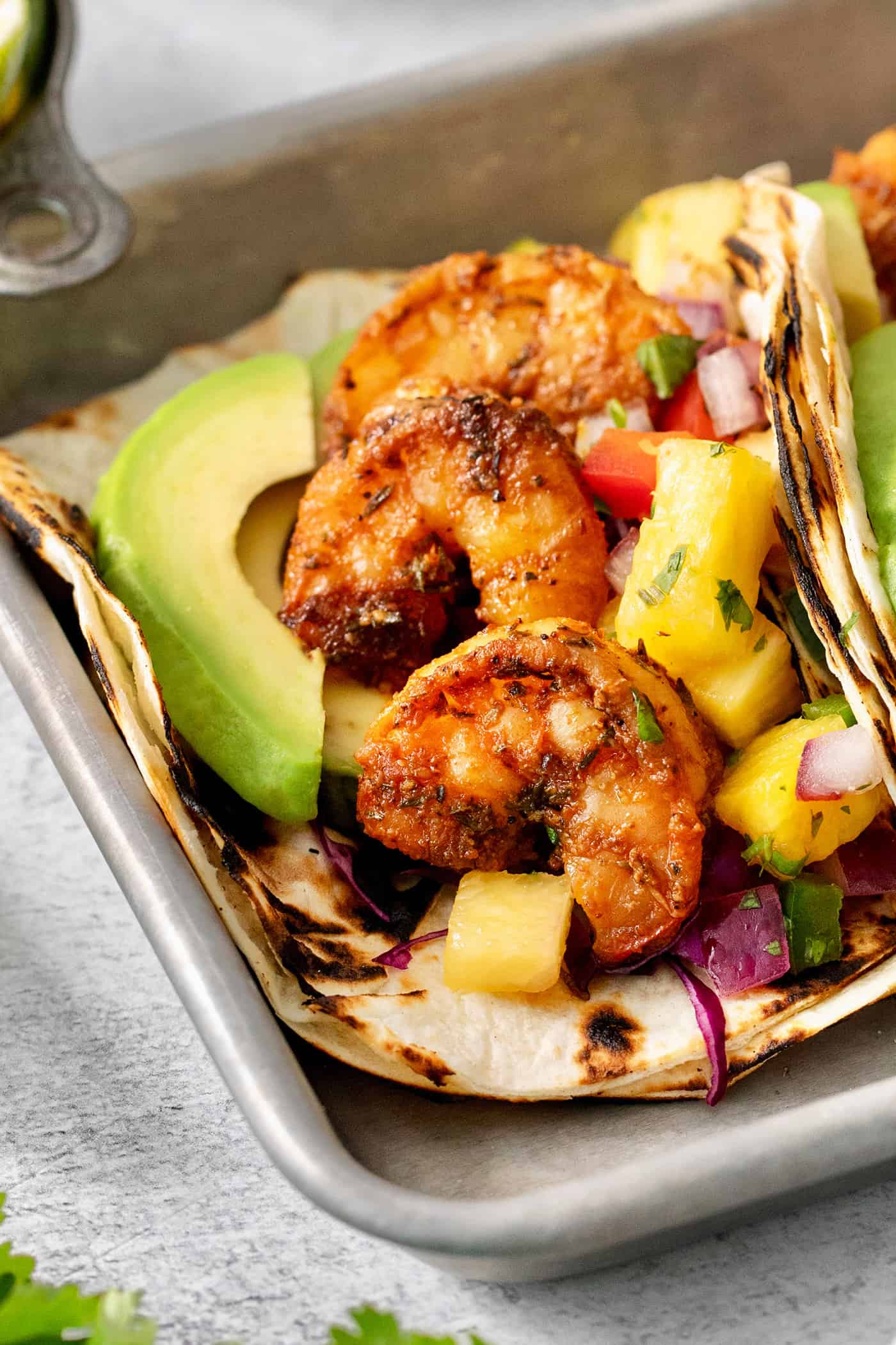 These blackened shrimp tacos feature charred flour tortillas stuffed with spicy blackened shrimp, pineapple salsa, and refreshing lime crema. A charred flour tortilla is opened to show blackened shrimp, an avocado slice, and pineapple salsa.