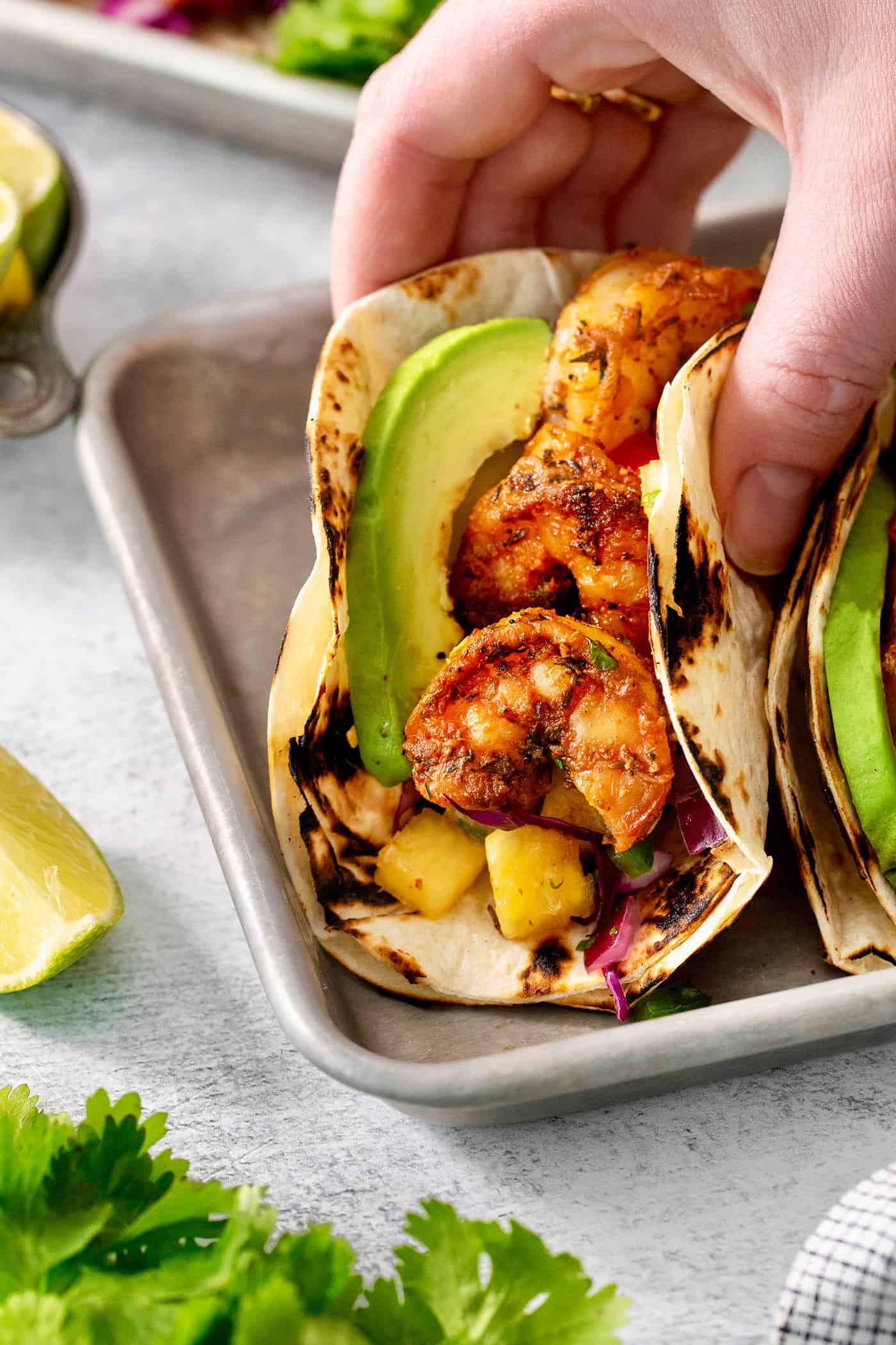 These blackened shrimp tacos feature charred flour tortillas stuffed with spicy blackened shrimp, pineapple salsa, and refreshing lime crema. A hand lifts up a perfectly assembled blackened shrimp taco.