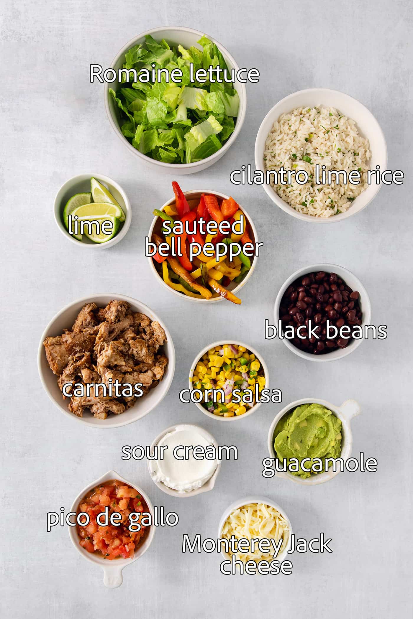 These Carnitas Burrito Bowls layer carnitas, black beans, citrusy cilantro lime rice, and pico de gallo for a better-than-takeout meal. The ingredients for carnitas burrito bowls are shown portioned out and labeled: carnitas, sweet red peppers, black beans, corn salsa, guacamole, pico de gallo, lime, cilantro rice, lettuce, jack cheese, sour cream.