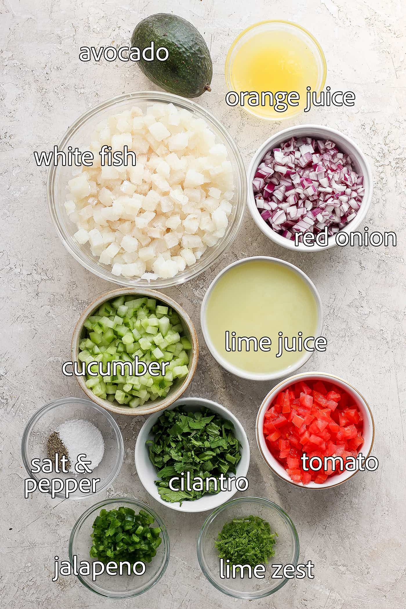 This refreshing Classic Ceviche recipe marinates fresh fish in lime and orange juices, then adds a mix of fresh veggies and a bit of jalapeno. The ingredients for classic ceviche are shown portioned out and labeled: white fish, lime juice, orange juice, tomato, red onion, cilantro, salt and pepper, cucumber, avocado.