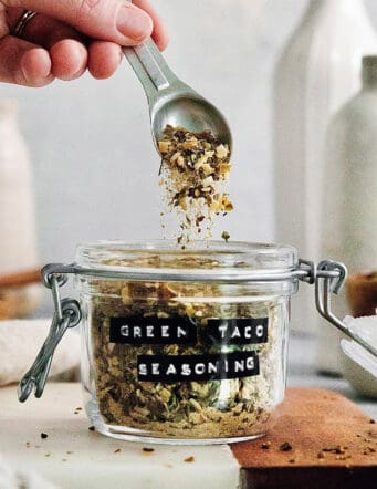 A hand holding a spoon that is dropping green taco seasoning into a jar.