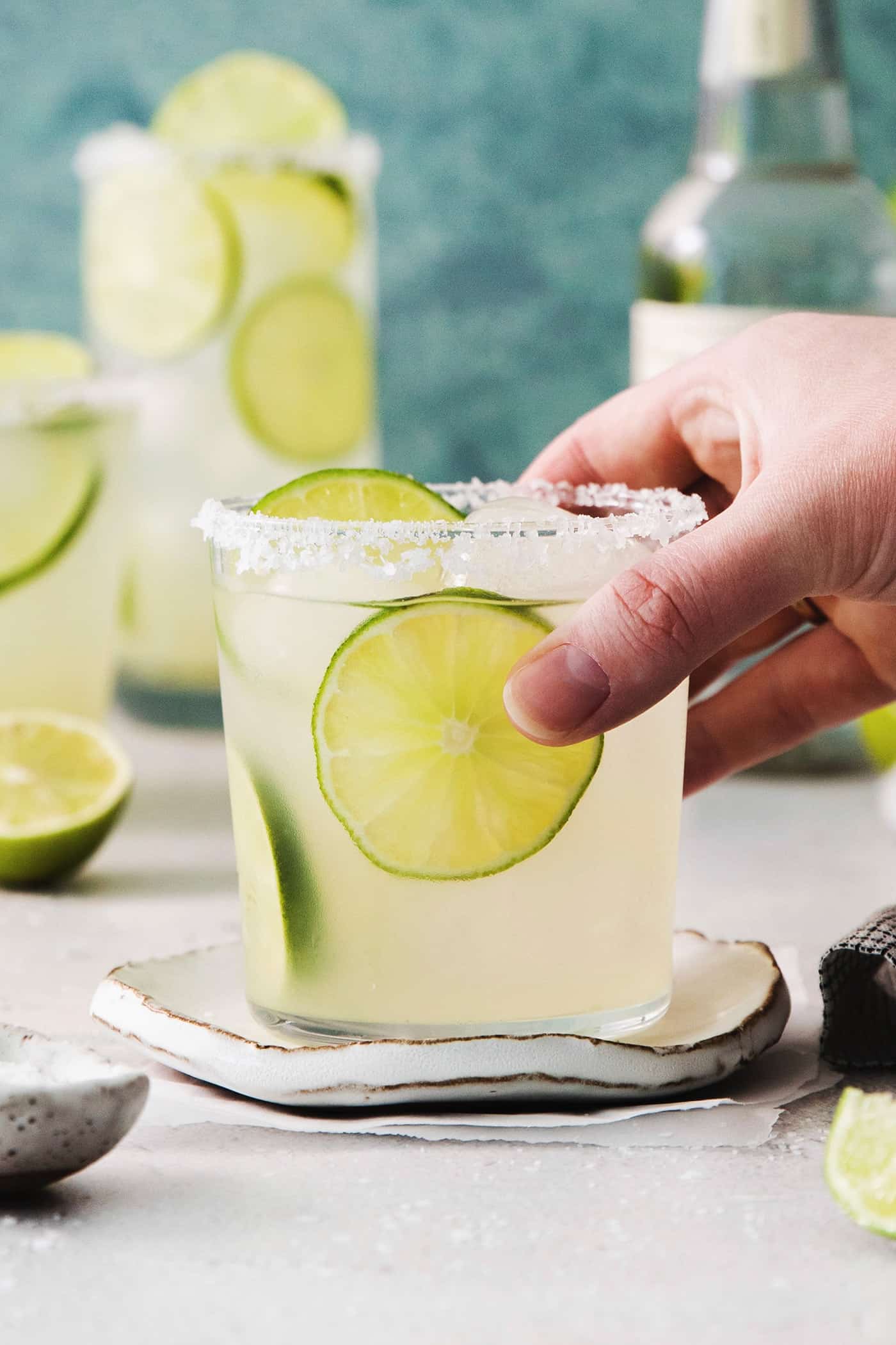 This Classic Margarita recipe makes the perfect tequila cocktail! Learn how to make a margarita that's always crisp, cold, and refreshing. A hand reaches to pick up a glass of classic margarita with more margaritas in the background.
